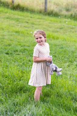 A smiling girl in a dress stands in the grass, with a stuffed animal clipart