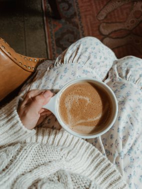 El ele tutuşup kahve fincanını çiçekli bir eteğin üzerine örülmüş bir kazak giyerek.