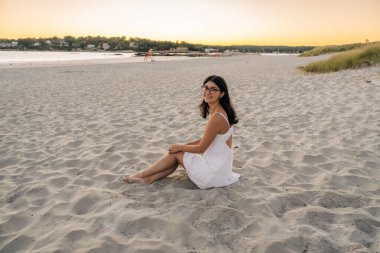 Gün batımında kumlu sahilde oturan beyaz elbiseli bir kadın.