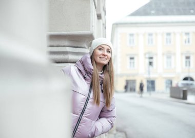 Açık hava binasına yaslanmış, kışlık ceketli, gülümseyen kadın.