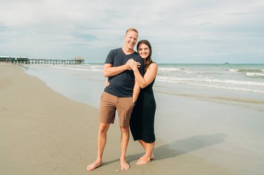 Bir çift, kumlu bir sahilde, rıhtımın yanında duruyor, sarılıyor ve gülümsüyor.