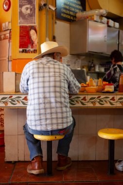 Grown man sitting on short stool at restaurant in Mexico clipart