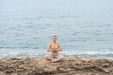 Genç bir kadın sahilde, dışarıda kayalarla yoga yapıyor.