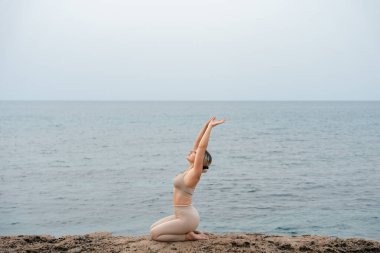 Genç bir kadın sahilde, dışarıda kayalarla yoga yapıyor.