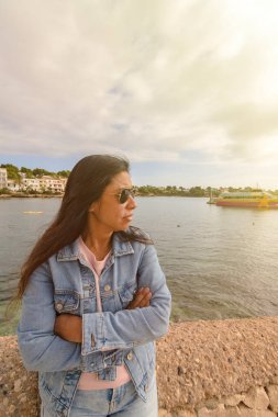 Kot ceketli ve güneş gözlüklü Latin kadın denize bakıyor.