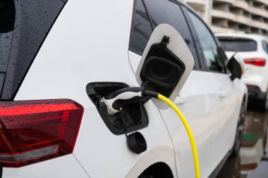 Sokak otoparkında beyaz elektrikli araba şarj oluyor.