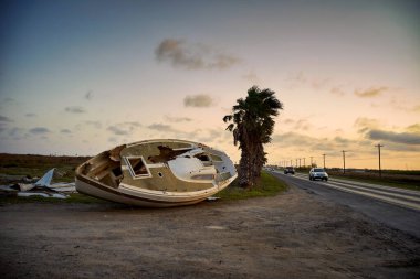 A boat lies overturned beside a road as vehicles pass by. clipart