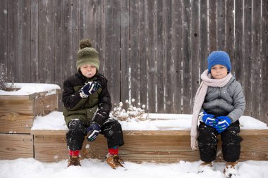 Two children bundled in winter clothing sitting while one eats snow clipart