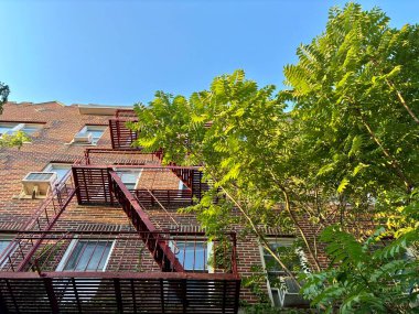 Yangın çıkışlı kırmızı tuğlalı apartman, yeşil ağaçlar, mavi gökyüzü.