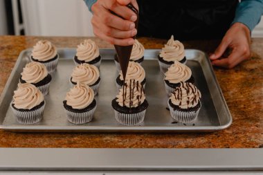 Female baker drizzling chocolate sauce on cupcakes on tray clipart