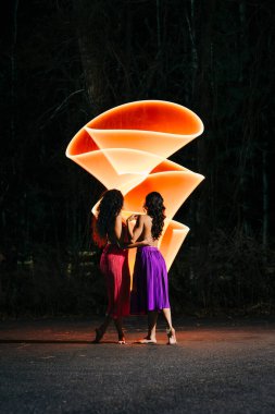 Two women in colorful skirts with glowing orange light trails at night clipart