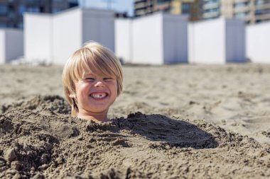 Kumsalda sırıtan neşeli bir sarışın çocuk..