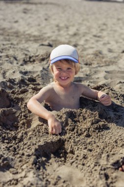 Şapka takan ve kuma gömülü bir çocuk kameraya bakıyor.