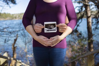 Soon to be parents pose with ultrasound photo in front of mother clipart