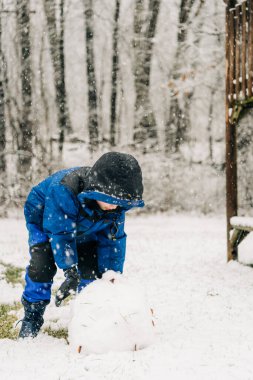 Mavi ceketli çocuk karlı bahçede ağaçlarla büyük bir kartopu şekillendiriyor.