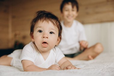 Bebek yatakta yatıyor, arkasında ağabeyi oturuyor.
