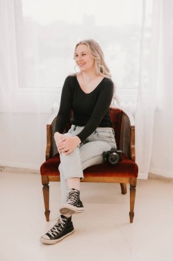 Kırmızı bir sandalyede otururken elinde kamerayla gülümseyen kadın.