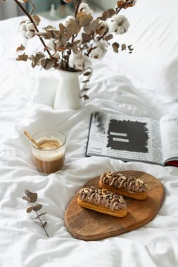 Ekler, kahve, açık kitap, kurumuş çiçekler. Yatakta kahvaltı.