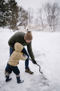 Bir kış fırtınasından sonra oğluyla birlikte garaj yolunda kar kürüyen bir adam.