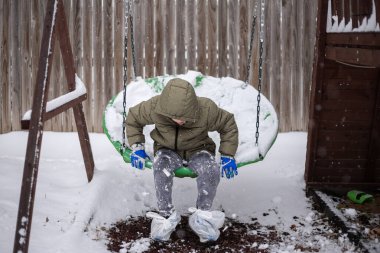 Çocuk kar kaplı bir salıncakta oturuyor, ayakkabılarında plastik torbalarla.