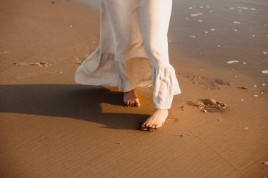 Gün batımında dalgalı beyaz elbiseli bir kadın okyanus dalgalarında duruyor.