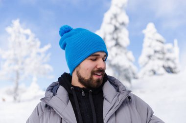 Karla kaplı ağaçlara ve açık mavi gökyüzüne karşı şık sakallı adam
