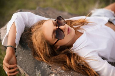 Kaya üzerinde güneş gözlüklü, rahat, genç, kızıl bir kadın.