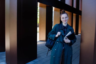 Gözlüklü, sırt çantalı ve modern binanın yanında kitapları olan kızıl saçlı kız.