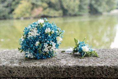 Mavi ve beyaz gelin buketi ve bir gölün kenarındaki bankta yaka çiçeği