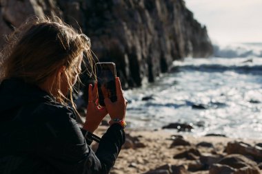 Kadın akıllı telefonuyla okyanus dalgalarını ve uçurumları fotoğraflıyor.