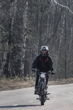 15 yaşında, kasklı bir çocuk kırsal yolda motosikletini sürüyor.