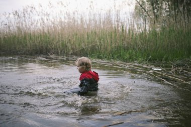 Küçük çocuk bulutlu bir günde sazlıkların yanında gölde su sıçratıyor.