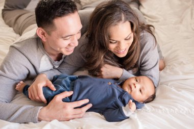 Gülümseyen anne babalar, küçük oğlana sarılır, rahat beyaz yatakta.