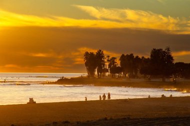 Palmiye ağaçlarının üzerinde altın bir günbatımı. İnsanlar sahilde eğleniyor.
