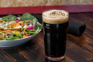 Bir bardak koyu bira, üzerinde köpüklü bir kafa ve salata.