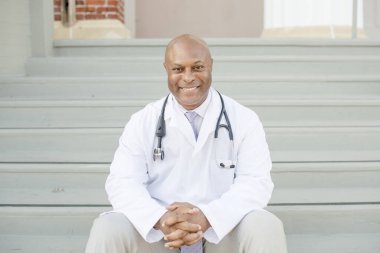 Dışarıdaki merdivenlerde oturan beyaz önlüklü gülümseyen doktor.