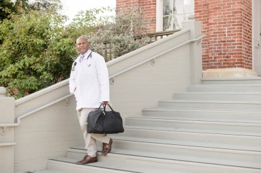 Beyaz önlüklü doktor elinde sağlık çantasıyla merdivenlerden iniyor.