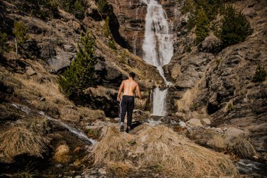 Tişörtsüz bir adam dağ şelalesine doğru yürüyor.