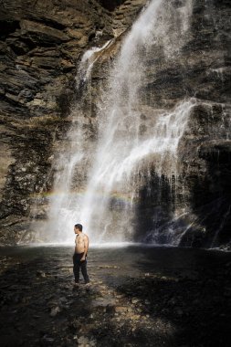 Gömleksiz bir adam dağ şelalesi gökkuşağının yanında