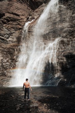 Gökkuşağı görünen bir şelalenin yanında duran adam.