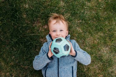 Gülen çocuk çimenlerde uzanmış elinde bir futbol topu tutuyor..