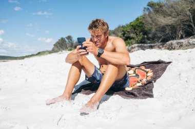 Plaj havlusunda oturan adam Jervis Körfezi sahil şeridinde telefon kullanıyor.