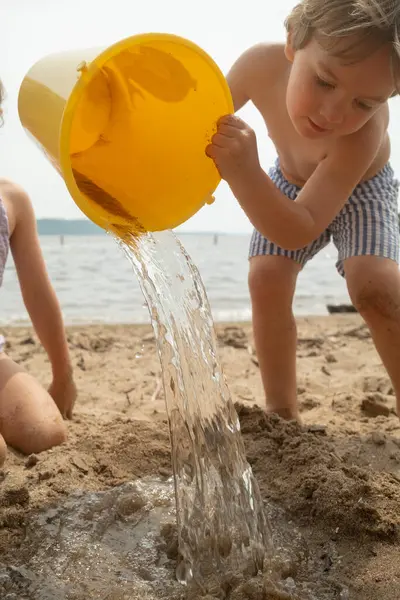 Çocuk sarı kovadan kumsala su döker..