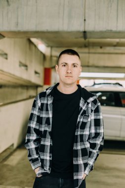 Ekose gömlekli genç adam otoparkta duruyor.