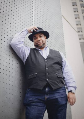 Kot pantolonlu siyah bir adam şapkasını gri bir duvara değdirir.