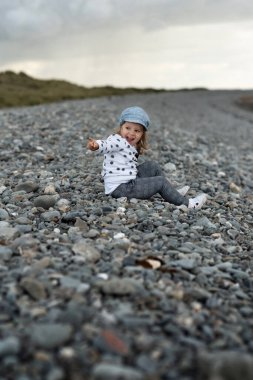 Mutlu küçük kız taş plajda oturuyor, gülüyor ve işaret ediyor.