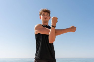 Genç sporcu mavi bir gökyüzüyle kumsalda kollarını açıyor.