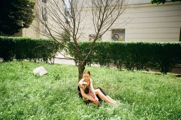 Çimenlerde oturan bir kadın kurak bir ağacın yanında köpeğine sarılıyor.