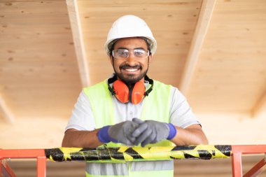 Beyaz miğferi ve güvenlik yeleği olan işçi ahşap bir ev inşa ediyor.