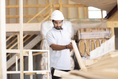 Beyaz miğferi ve güvenlik yeleği olan işçi ahşap bir ev inşa ediyor.